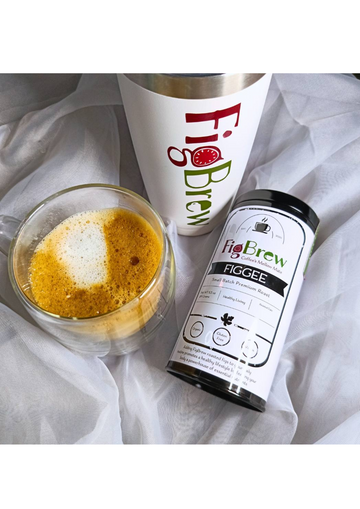 An image showcasing a FigBrew product. There is a clear glass cup filled with a frothy beverage, a FigBrew tumbler, and a canister labeled 'Figgee' from FigBrew. The items are arranged on a light-colored fabric backdrop.